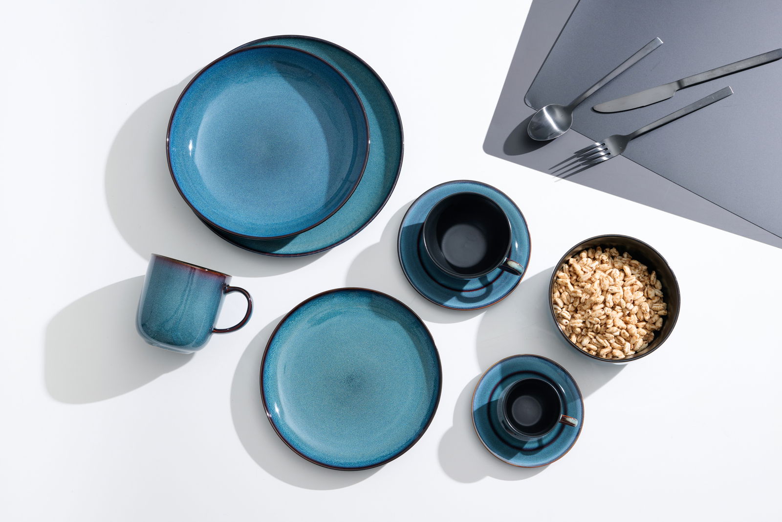 Vogelperspektive auf ein 8-teiliges Tafelservice in Blau- und Grüntönen, bestehend aus Tellern, Tassen und Besteck, auf einem weißen Hintergrund.