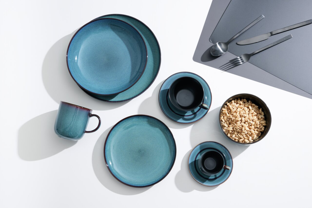 Vogelvluchtperspectief op een 8-delig servies in blauw- en groentinten, bestaande uit borden, kopjes en bestek, op een witte achtergrond.