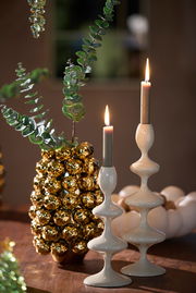 Zwei elegante, weiße Kerzenständer mit geschwungenem Design, auf einem Tisch arrangiert, mit brennenden Kerzen und dekorativen Elementen im Hintergrund. Seitliche Perspektive.