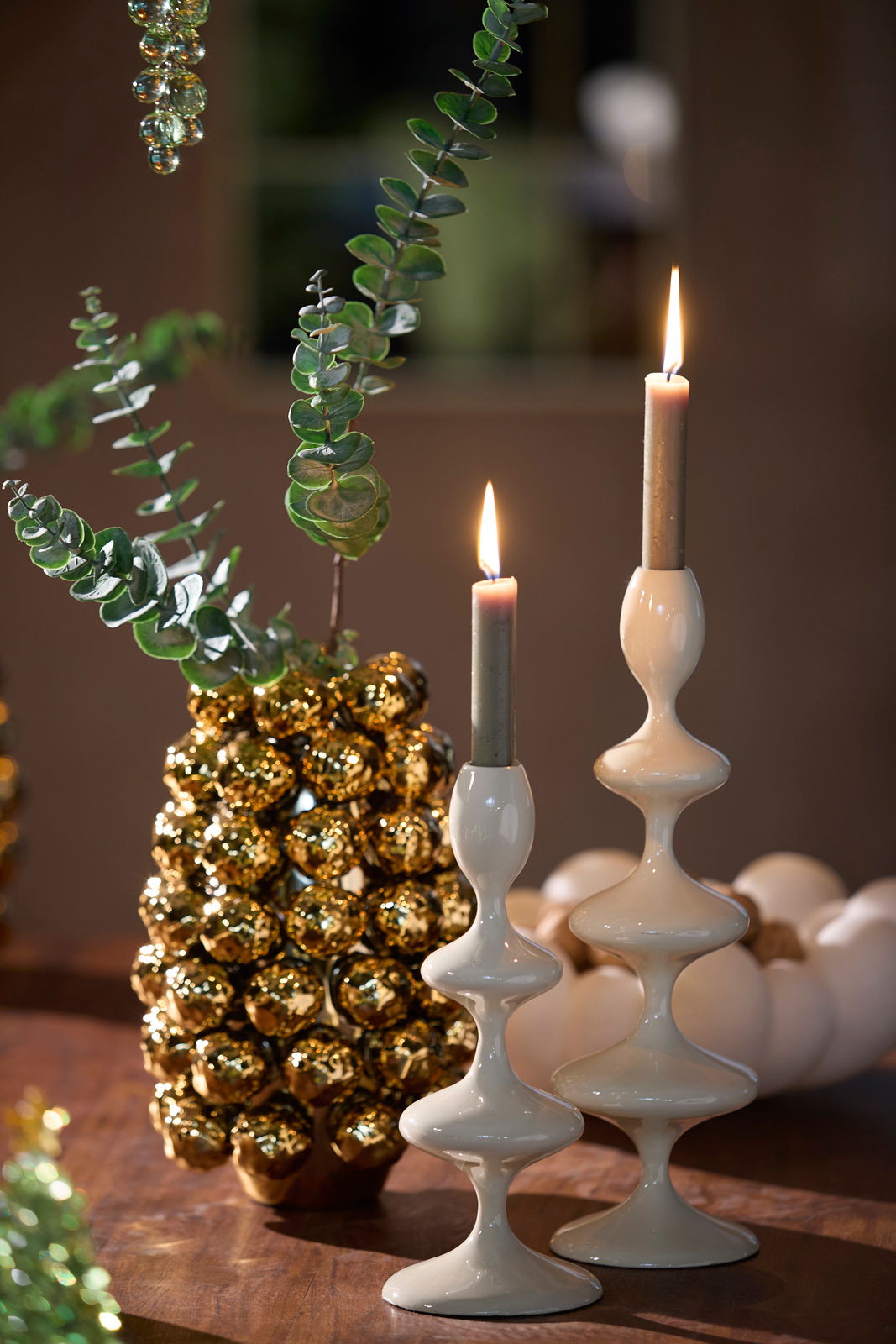 Zwei elegante, weiße Kerzenständer mit geschwungenem Design, auf einem Tisch arrangiert, mit brennenden Kerzen und dekorativen Elementen im Hintergrund. Seitliche Perspektive.