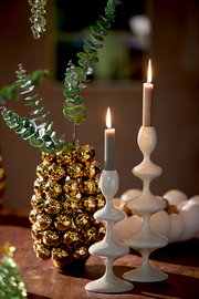 Zwei elegante, weiße Kerzenständer mit geschwungenem Design, auf einem Tisch arrangiert, mit brennenden Kerzen und dekorativen Elementen im Hintergrund. Seitliche Perspektive.