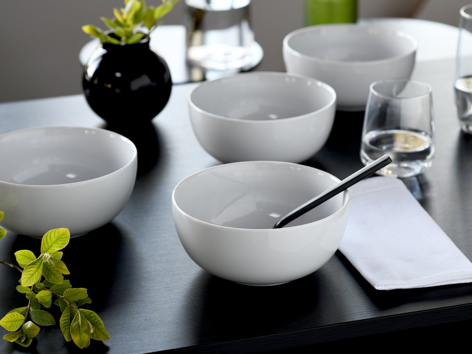 Weiße, minimalistische Keramikschalen auf dunklem Tisch mit Wassergläsern, schwarzer Vase und gefaltetem weißen Tuch; Perspektive: leicht von oben in schräger Ansicht.