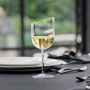 Ein eleganter Weißweinkelch mit modernem, schrägem Rand, gefüllt mit Weißwein, auf einem gedeckten Tisch aus seitlicher Perspektive.