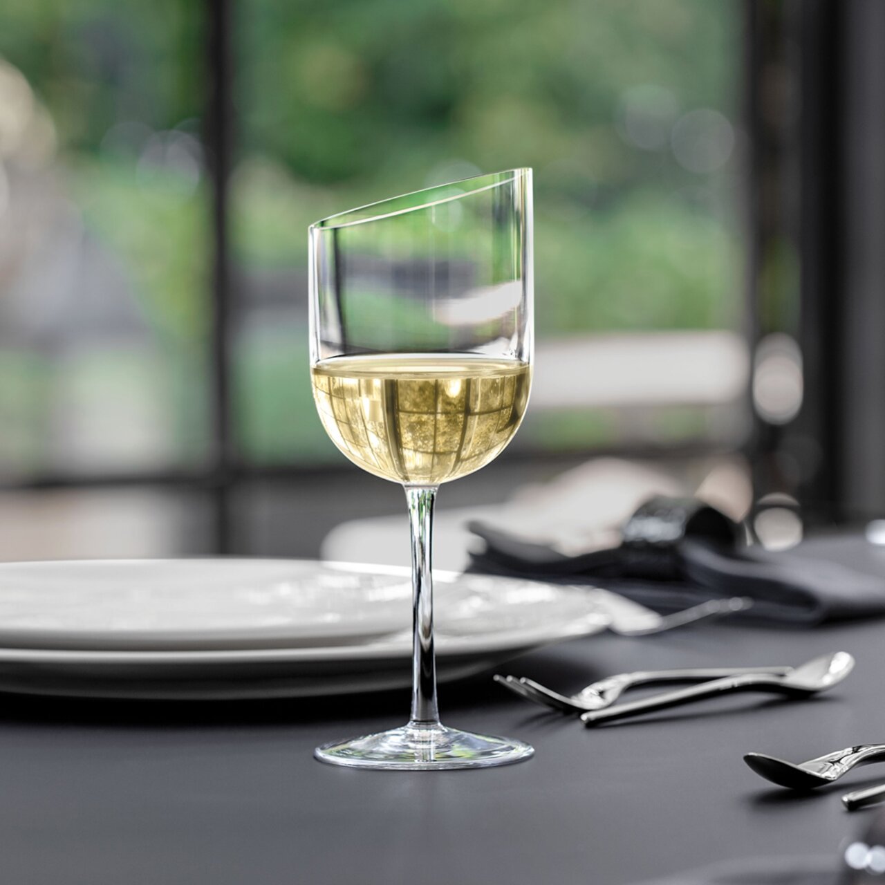 Ein eleganter Weißweinkelch mit modernem, schrägem Rand, gefüllt mit Weißwein, auf einem gedeckten Tisch aus seitlicher Perspektive.
