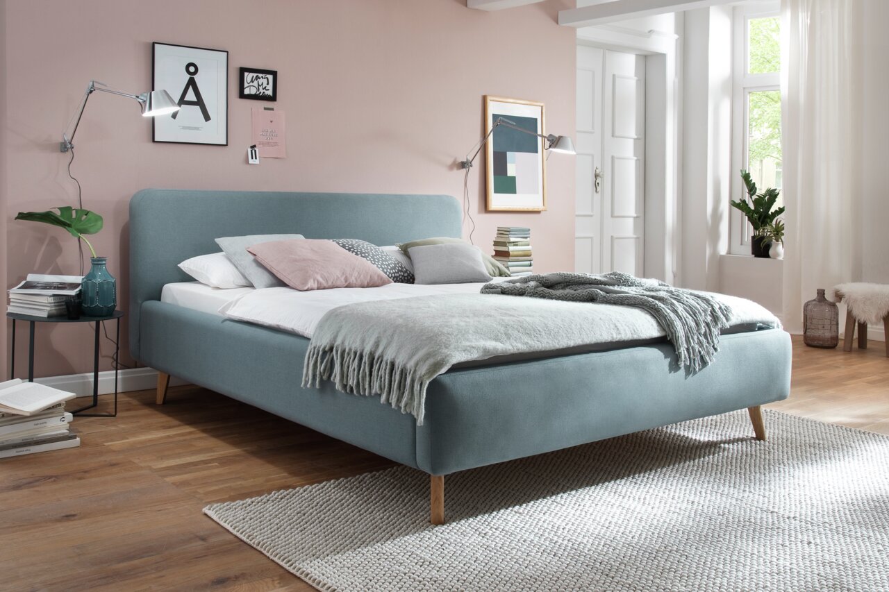 Z2 Polsterbett MATTIS Polsterbett in einem modernen Schlafzimmer, seitliche Perspektive, mit blauer Stoffpolsterung und Holzfüßen, dekoriert mit Kissen und Decken.