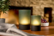 Zwei Akku-Windlichter in verschiedenen Größen auf einem Tisch, umgeben von einer Pflanze und einer Schale mit Trauben, aus seitlicher Perspektive fotografiert.