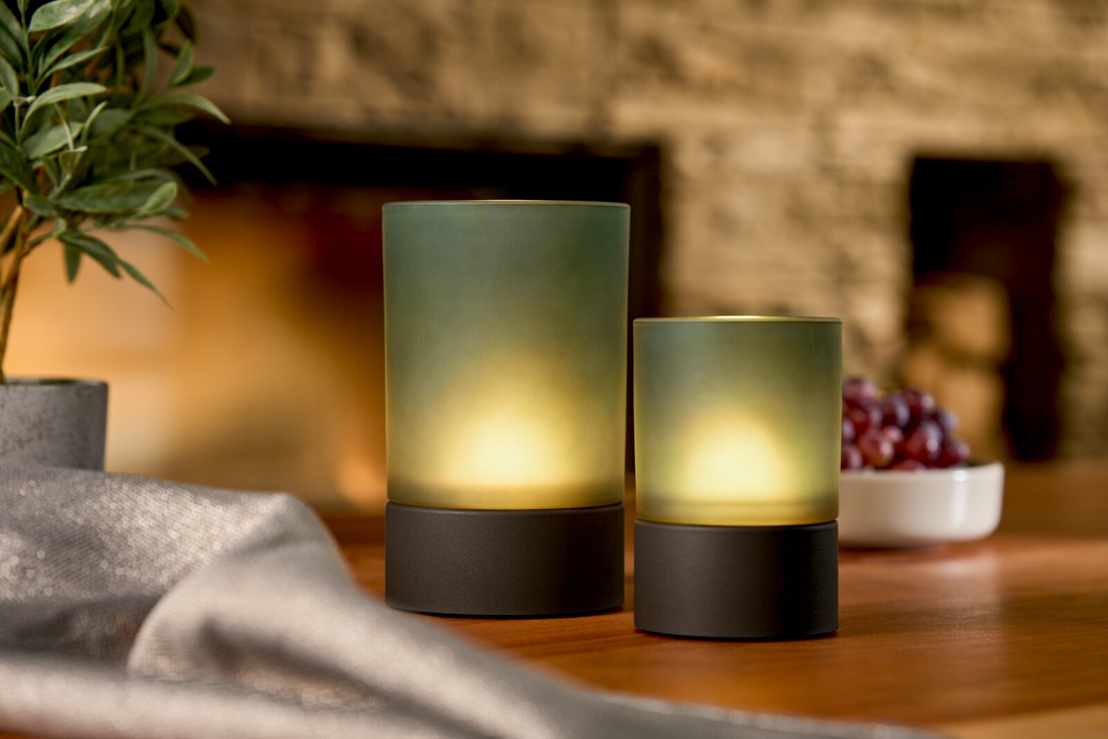 Zwei Akku-Windlichter in verschiedenen Größen auf einem Tisch, umgeben von einer Pflanze und einer Schale mit Trauben, aus seitlicher Perspektive fotografiert.