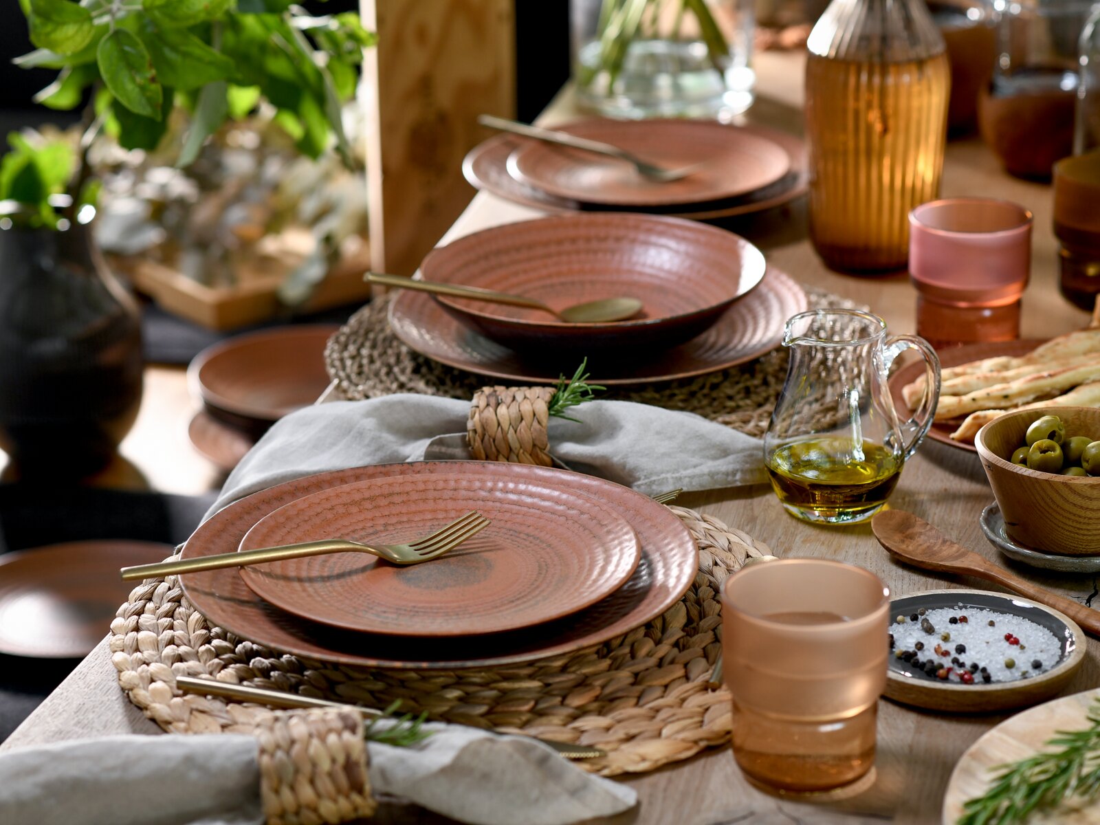 Rustikales Tischgedeck mit roséfarbenem Keramikgeschirr auf geflochtenen Platzsets, goldfarbenem Besteck, Glasflasche und Olivenölkaraffe, aus leicht schräger Seitenansicht