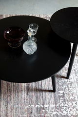 Zwei schwarze runde Couchtische aus der Vogelperspektive, dekoriert mit einer Kristallkaraffe, einem Weinglas und einer dunklen Glasschale, auf einem gemusterten Teppich.