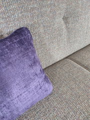 Nahaufnahme in schräger Seitenperspektive: graues, strukturiertes Sofa mit lila Samt-Zierkissen auf der Sitzfläche.