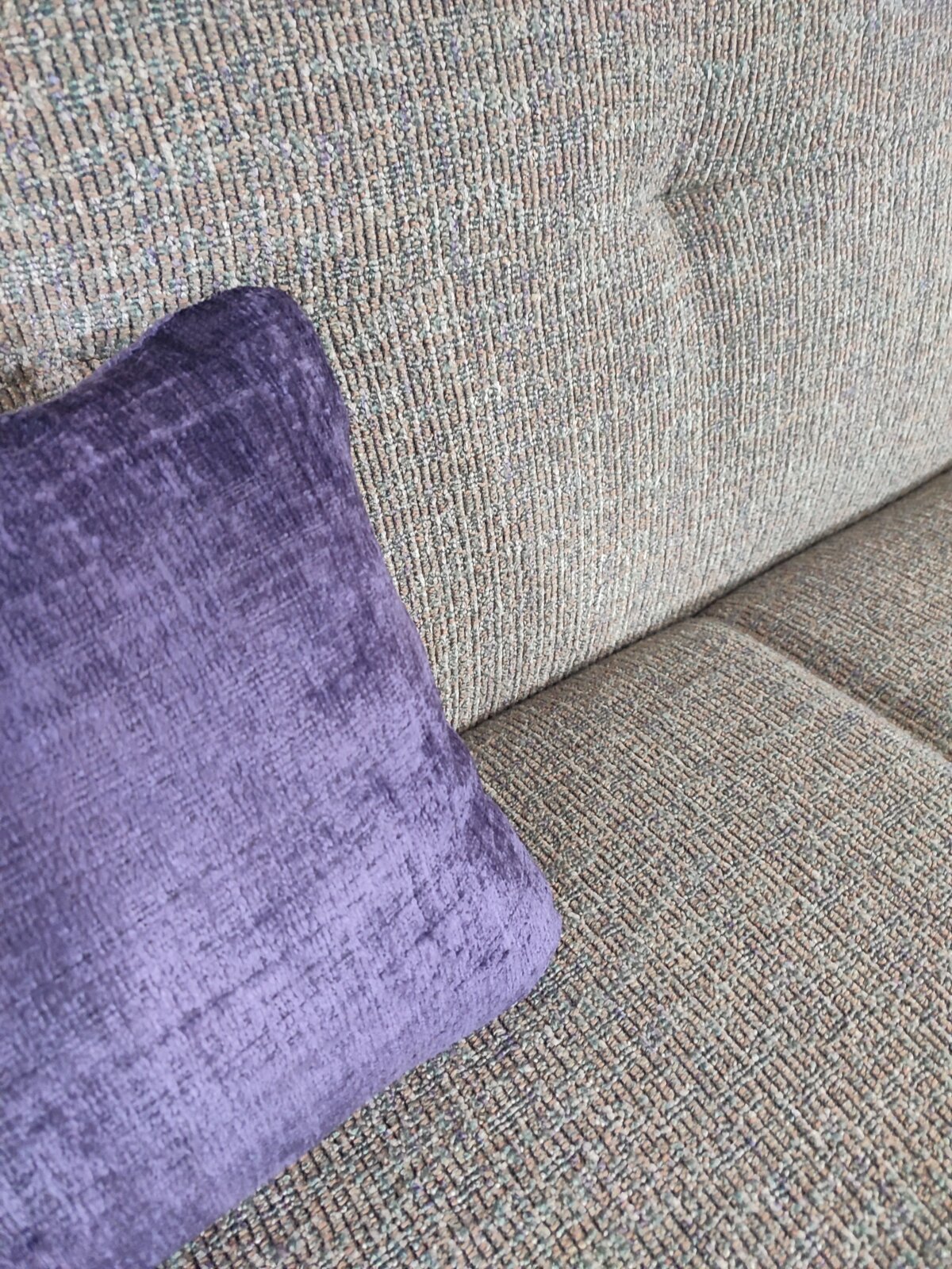 Nahaufnahme in schräger Seitenperspektive: graues, strukturiertes Sofa mit lila Samt-Zierkissen auf der Sitzfläche.
