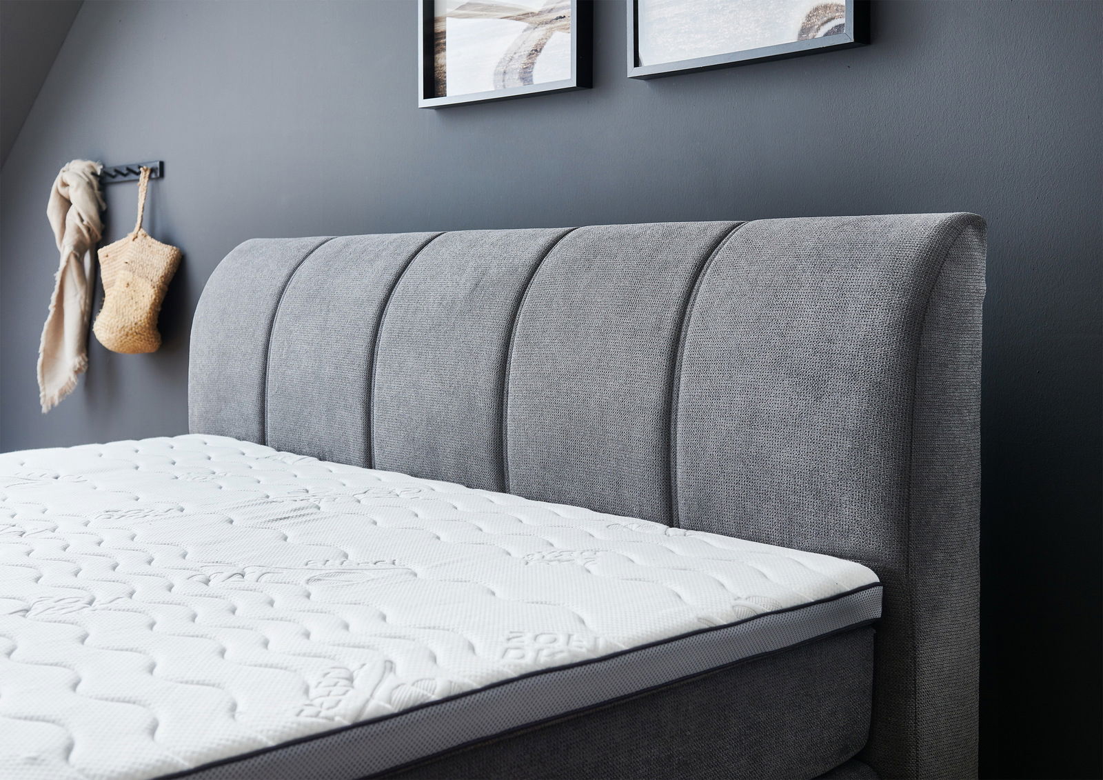 Seitliche Ansicht eines grauen Boxspringbetts mit gepolstertem Kopfteil und weißer Matratze in einem modern eingerichteten Raum.