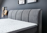 Seitliche Ansicht eines grauen Boxspringbetts mit gepolstertem Kopfteil und weißer Matratze in einem modern eingerichteten Raum.