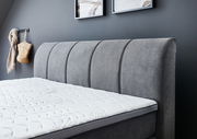 Seitliche Ansicht eines grauen Boxspringbetts mit gepolstertem Kopfteil und weißer Matratze in einem modern eingerichteten Raum.