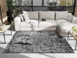 Moderner Wohnzimmerteppich in Grautönen, ca. 140x200cm, vor einem beigen Sofa in einem hellen Raum mit großen Fenstern, von oben betrachtet.