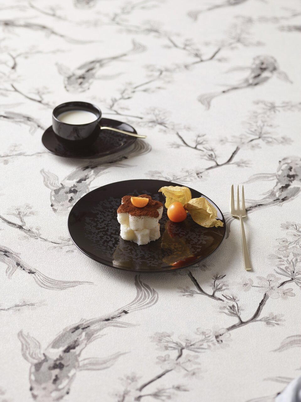 Schwarzes Tellerset Eclipse Night mit Dessert und Physalis, von oben fotografiert, auf einem Tisch mit floralem Muster.