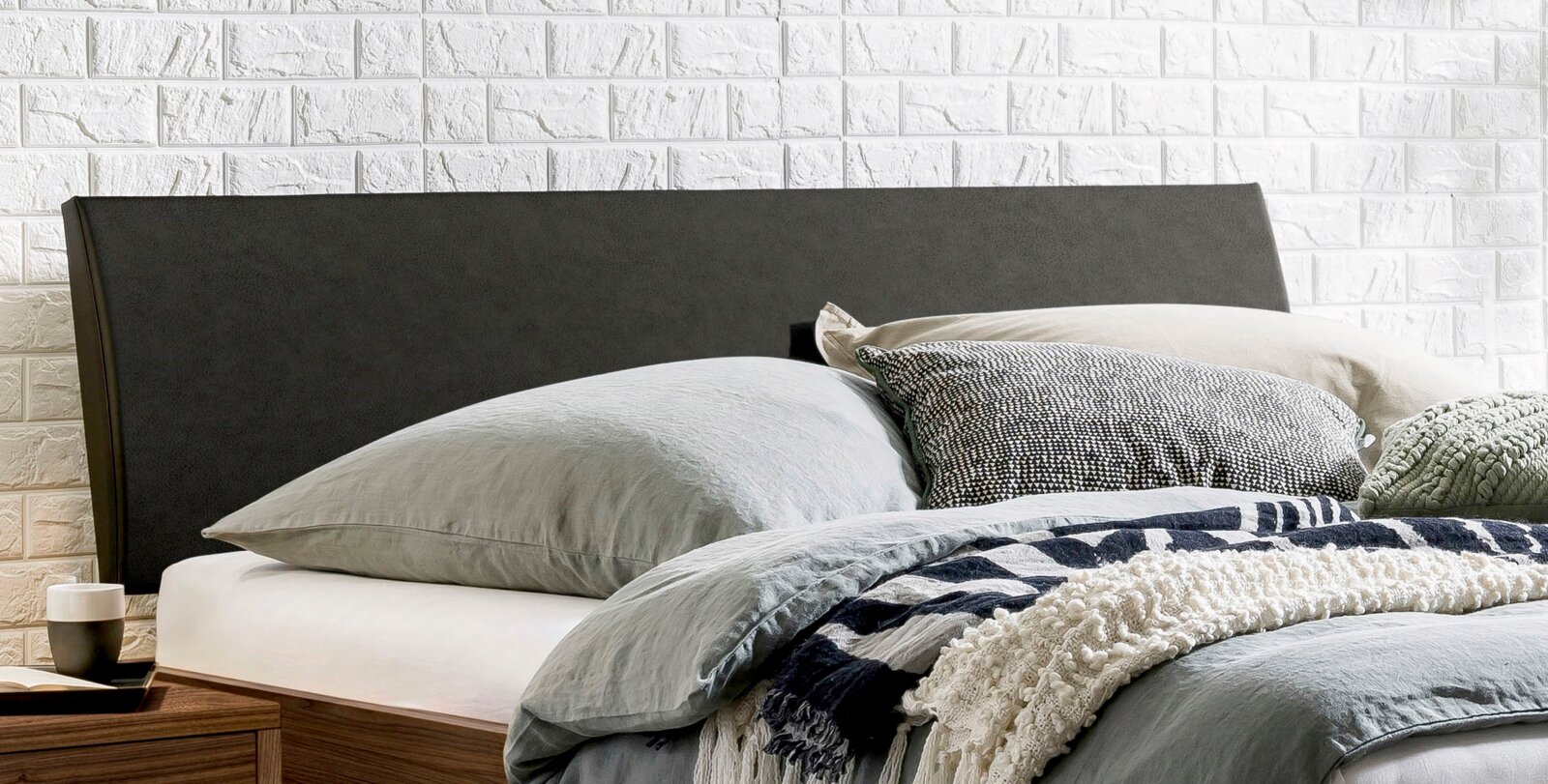Modern bed met een donkergrijs, gestoffeerd hoofdeinde en grijs beddengoed tegen een witte bakstenen muur, zijaanzicht