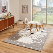 Kinderzimmer mit einem Merinos Kinderteppich LÖWENHERZ, der einen stilisierten Löwen in Grau- und Weißtönen zeigt, umgeben von einem Tisch und zwei Stühlen, mit Blick auf einen sonnigen Garten durch eine große Fensterfront.