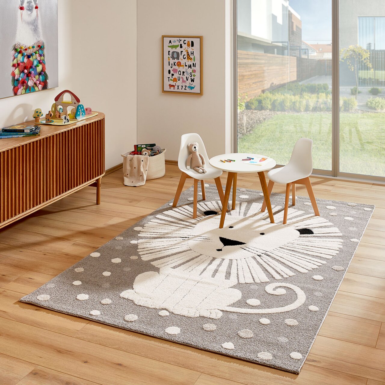 Kinderzimmer mit einem Merinos Kinderteppich LÖWENHERZ, der einen stilisierten Löwen in Grau- und Weißtönen zeigt, umgeben von einem Tisch und zwei Stühlen, mit Blick auf einen sonnigen Garten durch eine große Fensterfront.