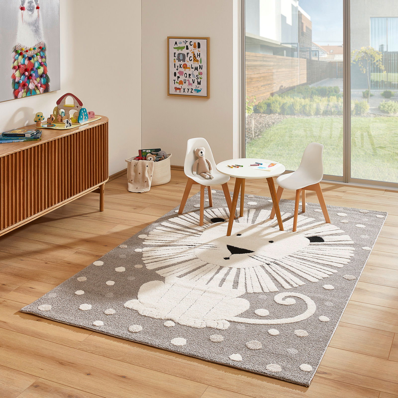 Kinderzimmer mit einem Merinos Kinderteppich LÖWENHERZ, der einen stilisierten Löwen in Grau- und Weißtönen zeigt, umgeben von einem Tisch und zwei Stühlen, mit Blick auf einen sonnigen Garten durch eine große Fensterfront.