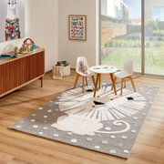 Kinderzimmer mit einem Merinos Kinderteppich LÖWENHERZ, der einen stilisierten Löwen in Grau- und Weißtönen zeigt, umgeben von einem Tisch und zwei Stühlen, mit Blick auf einen sonnigen Garten durch eine große Fensterfront.