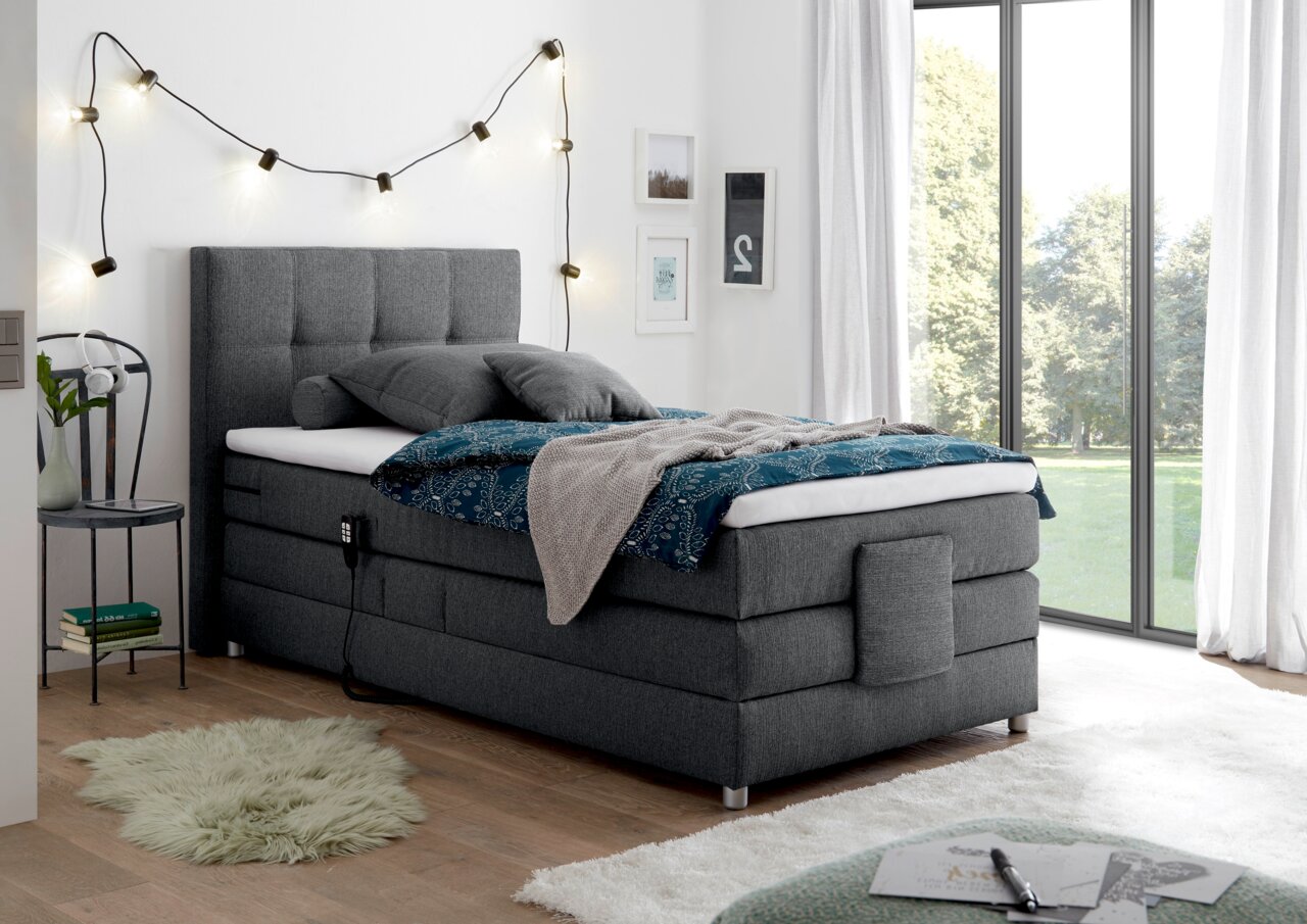 Graues Boxspringbett in einem modernen Schlafzimmer, seitliche Perspektive, mit dekorativen Kissen und Decken, umgeben von stilvollen Lichterketten und großen Fenstern.