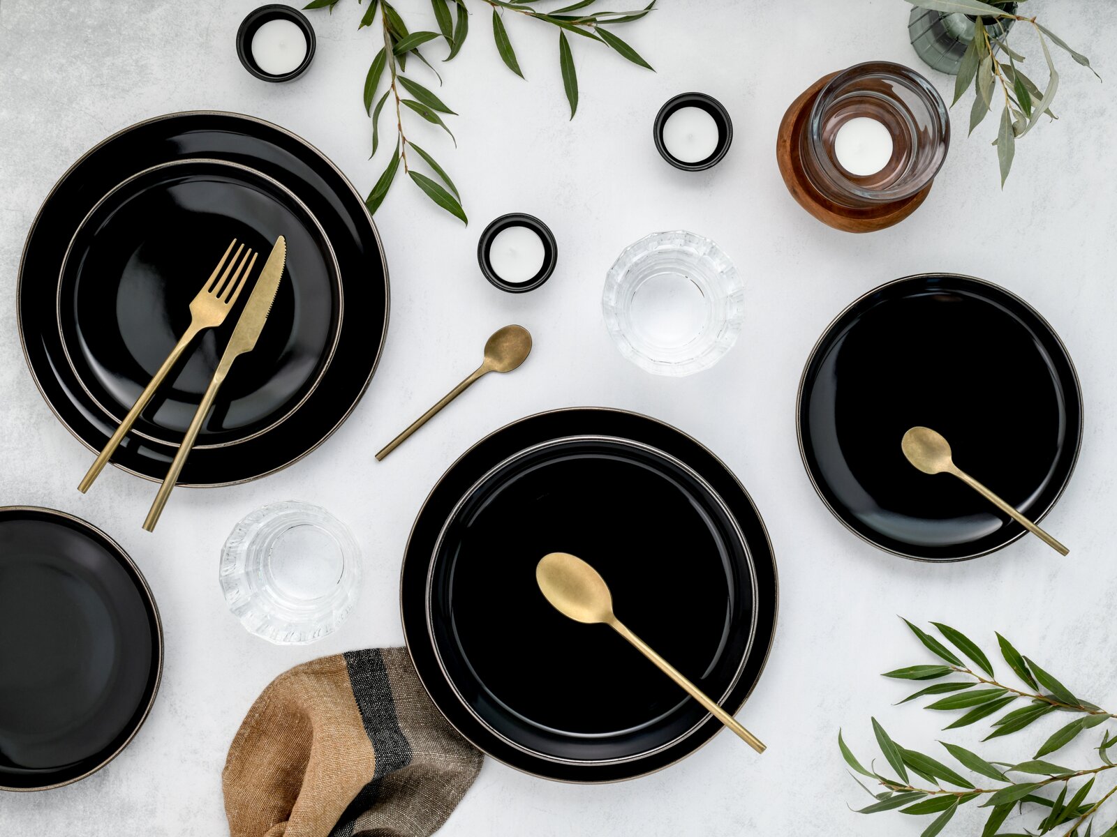 Draufsicht auf ein modern gedecktes Tisch-Arrangement mit schwarzen Tellern, goldfarbenem Besteck, Teelichtern, Wassergläsern und grünen Zweigen auf hellem Untergrund.