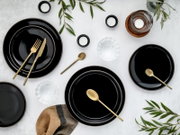 Draufsicht auf ein modern gedecktes Tisch-Arrangement mit schwarzen Tellern, goldfarbenem Besteck, Teelichtern, Wassergläsern und grünen Zweigen auf hellem Untergrund.