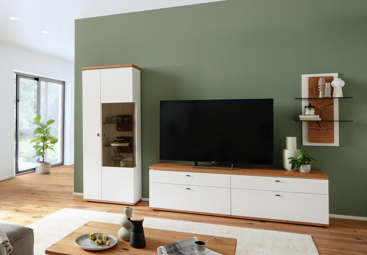Wandkombination MATADI in modernem Wohnzimmer, bestehend aus einem hohen Schrank mit Glastür, einem TV-Board und einem Wandregal, aus weißem Material mit Holzoberflächen, frontal betrachtet.