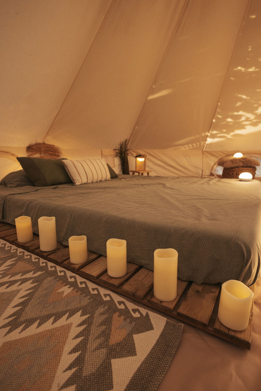 Ein gemütliches Zeltinterieur mit einem großen Bett, umgeben von mehreren Outdoor-Kerzen, die eine warme und einladende Atmosphäre schaffen. Die Perspektive zeigt das Bett von der Seite, mit den Kerzen im Vordergrund.