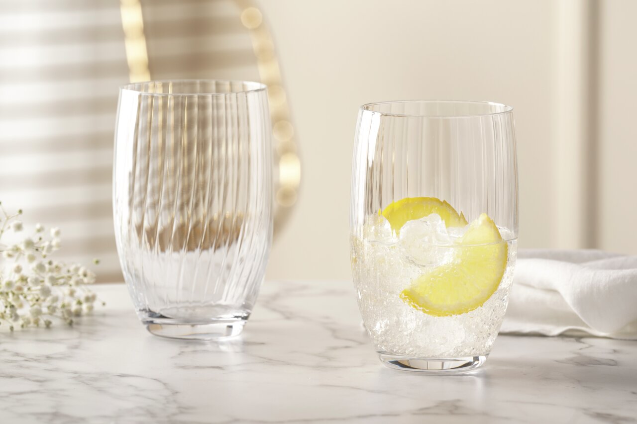 Zwei elegante Trinkgläser POESIA 460ml auf einem Marmortisch, eines gefüllt mit Eis und Zitronenscheiben, aus seitlicher Perspektive fotografiert.