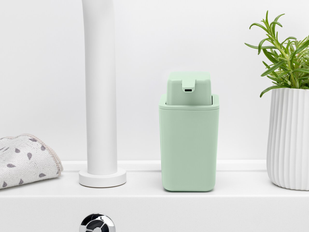 Een mintgroene zeepdispenser op een wastafel, van voren gefotografeerd, naast een handdoek en een plant.