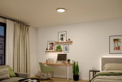 Runde, flache LED-Deckenleuchte in Weiß mit schmalem Rahmen, an der Zimmerdecke montiert, gezeigt in schräger Untersicht in einem modern eingerichteten Schlafzimmer/Homeoffice.