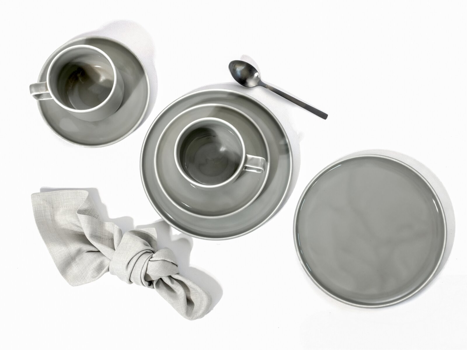Graues Keramik-Geschirrset mit zwei Tassen auf Untertassen, Teller, Löffel und Stoffserviette auf weißem Hintergrund, Draufsicht (Flatlay)