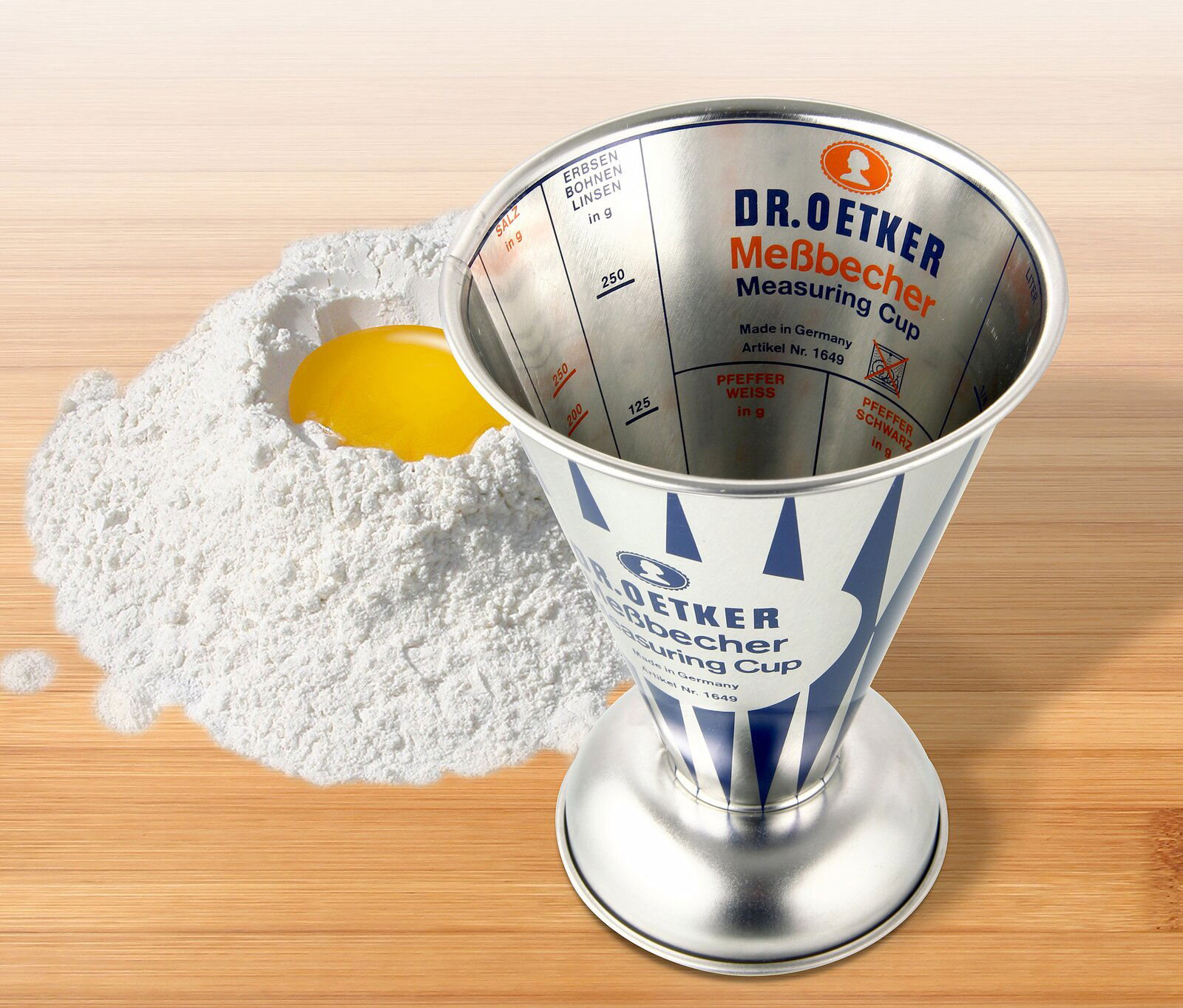 Dr. Oetker Messbecher 0,5L NOSTALGIE Nostalgischer 0,5L Messbecher von Dr. Oetker aus Metall, auf einer Holzoberfläche neben Mehl und einem aufgeschlagenen Ei, von oben betrachtet.