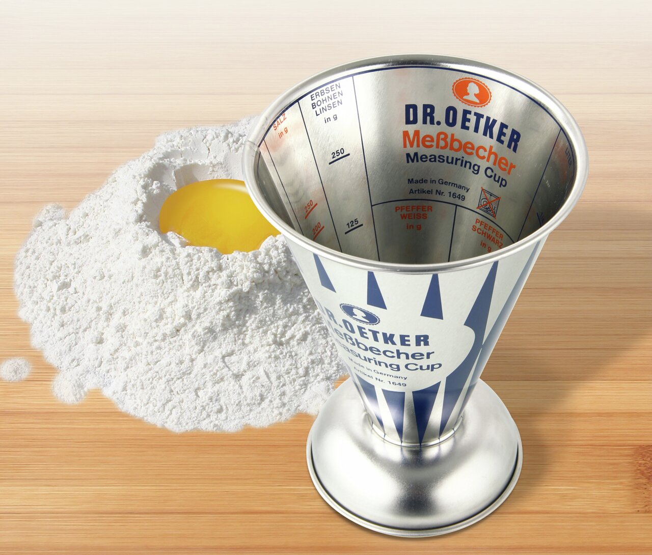 Nostalgischer 0,5L Messbecher von Dr. Oetker aus Metall, auf einer Holzoberfläche neben Mehl und einem aufgeschlagenen Ei, von oben betrachtet.