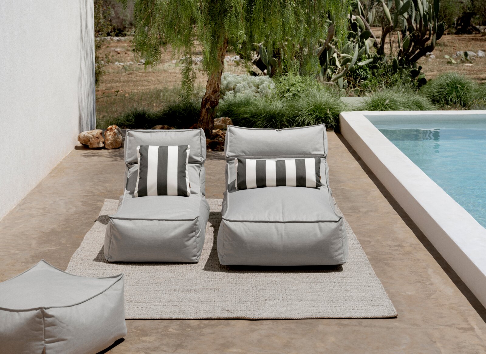 blomus Outdoor Hocker STAY Zwei graue Outdoor-Loungesessel/Sonnenliegen mit schwarz-weiß gestreiften Kissen auf hellem Teppich neben einem Pool, frontal in leichter Aufsicht fotografiert.