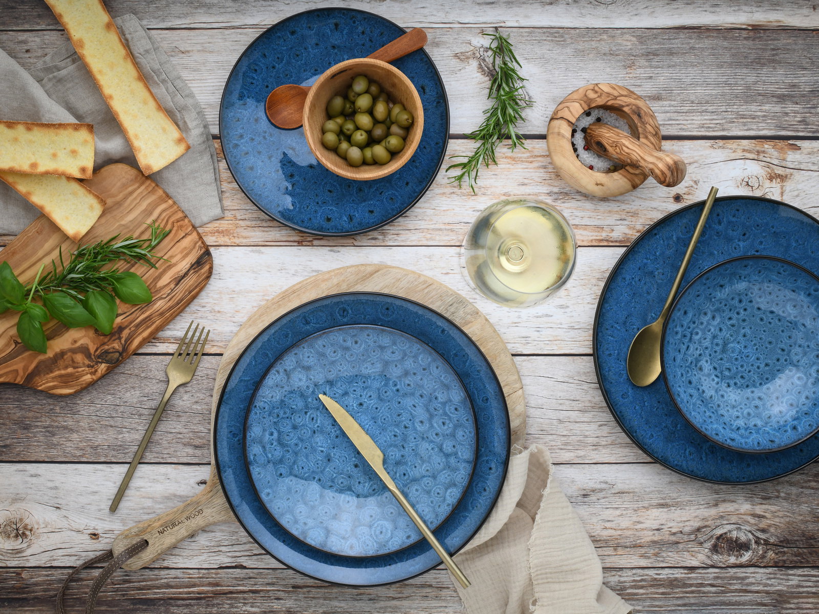 Vogelperspektive auf ein 18-teiliges Tellerset in Blau mit rustikalem Design, umgeben von Oliven, Kräutern, Brot und einem Glas Weißwein.