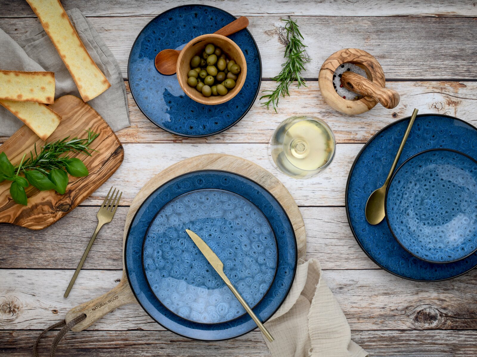 Vogelperspektive auf ein 18-teiliges Tellerset in Blau mit rustikalem Design, umgeben von Oliven, Kräutern, Brot und einem Glas Weißwein.