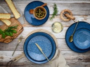 Vogelperspektive auf ein 18-teiliges Tellerset in Blau mit rustikalem Design, umgeben von Oliven, Kräutern, Brot und einem Glas Weißwein.