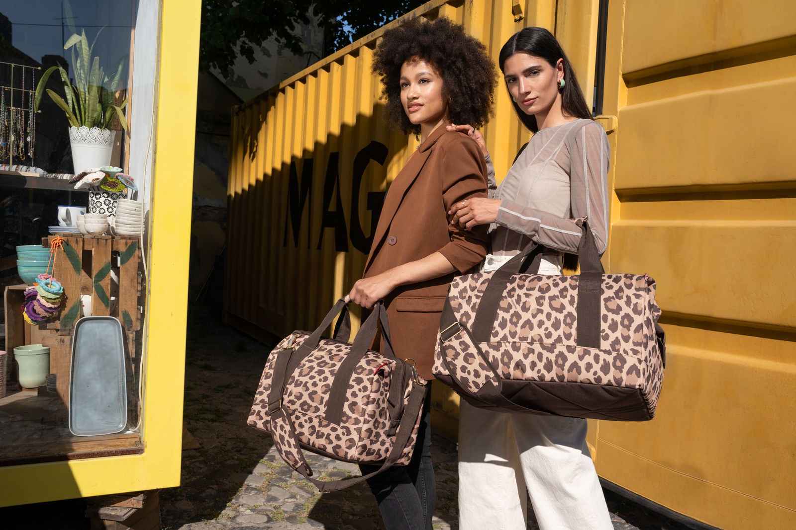 Zwei Frauen stehen vor einem gelben Container und halten Taschen mit Leopardenmuster. Die Taschen sind aus der Perspektive von vorne zu sehen.