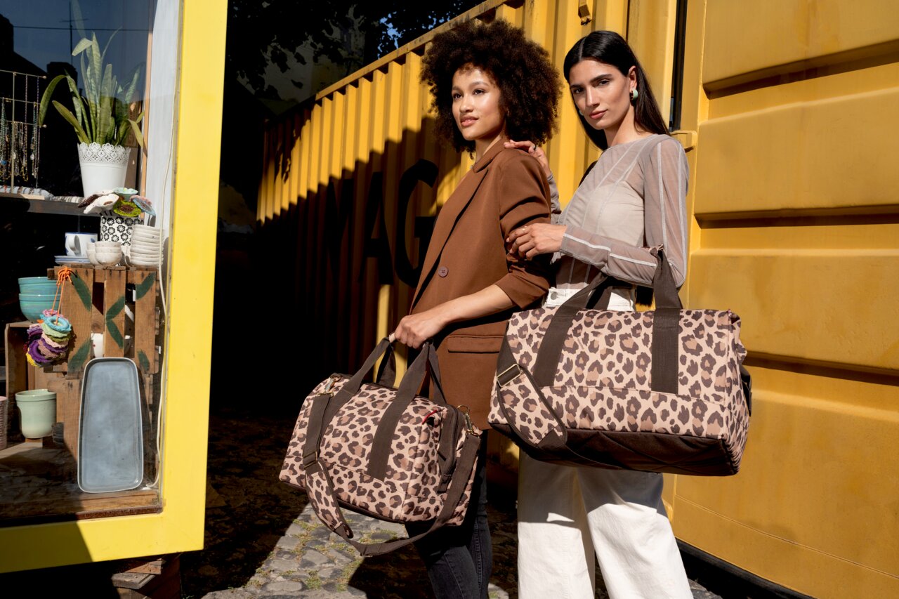 Twee vrouwen staan voor een gele container en houden tassen met luipaardprint vast. De tassen zijn van voren te zien.