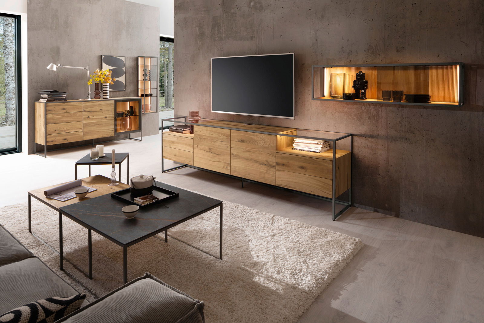 Modernes Wohnzimmer mit einem Venjakob Sideboard TAGENA aus Holz und Metall, frontal betrachtet. Das Sideboard steht unter einem großen Fernseher, umgeben von stilvollen Dekorationen und einem Hängeregal an der Wand.