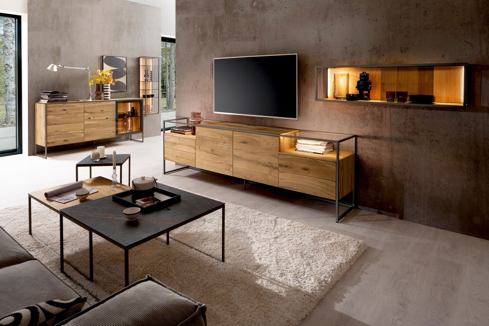 Modernes Wohnzimmer mit einem Venjakob Sideboard TAGENA aus Holz und Metall, frontal betrachtet. Das Sideboard steht unter einem großen Fernseher, umgeben von stilvollen Dekorationen und einem Hängeregal an der Wand.