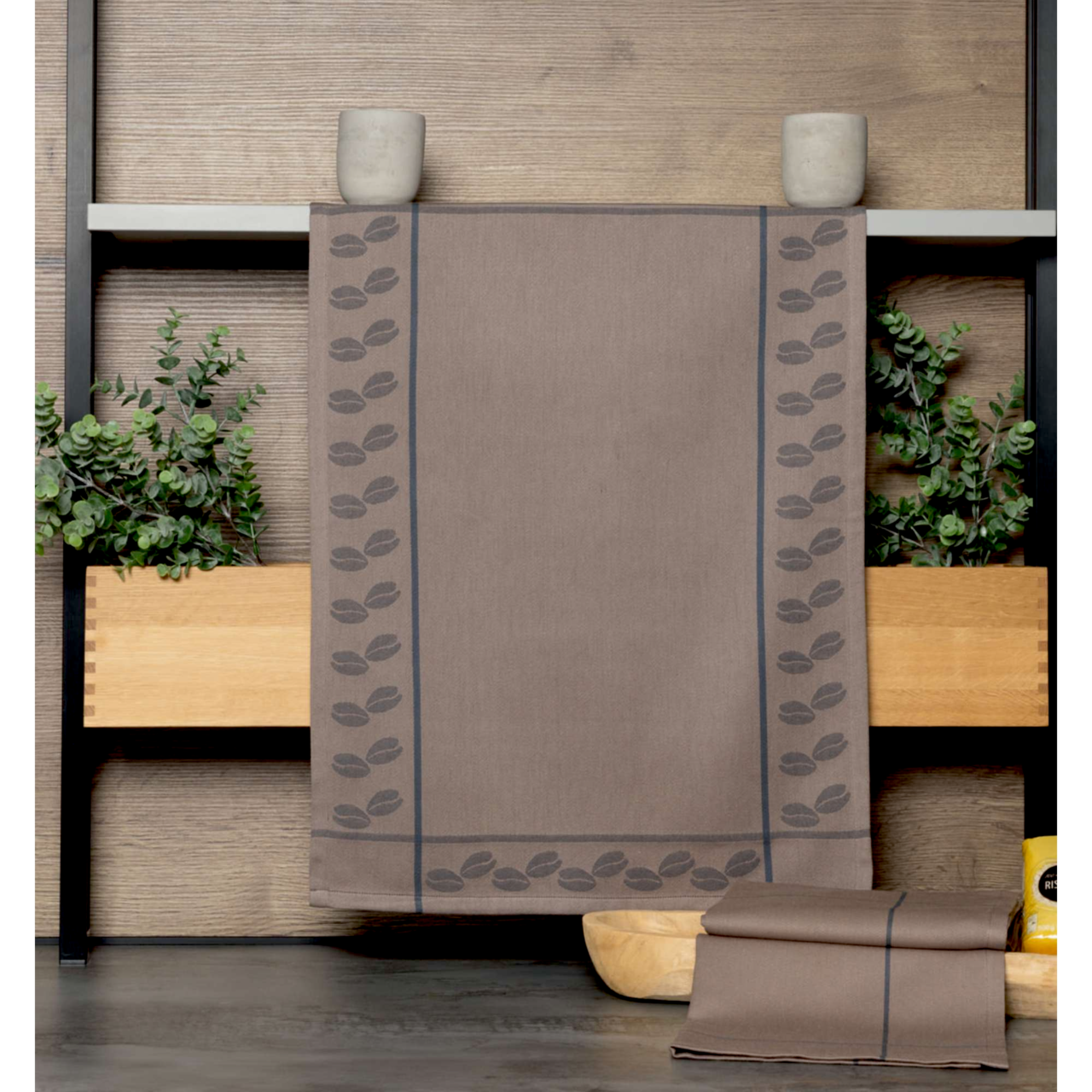 Geschirrtuch in Braun mit Kaffeebohnenmuster, frontal präsentiert auf einem Regal mit Pflanzen.