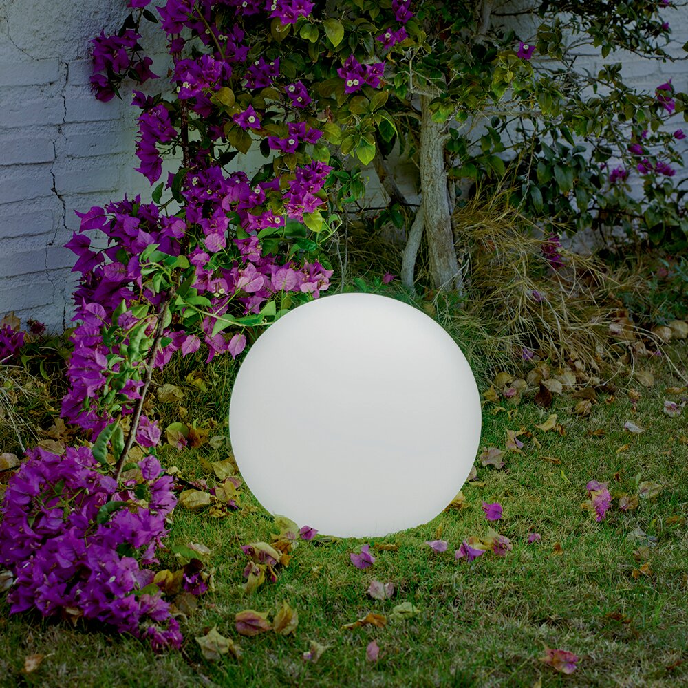 Leuchtende weiße Kugel-Gartenlampe auf dem Rasen vor einer Mauer, umgeben von violetten Bougainvillea-Blüten und Laub, aufgenommen in frontaler Perspektive auf Bodenhöhe.