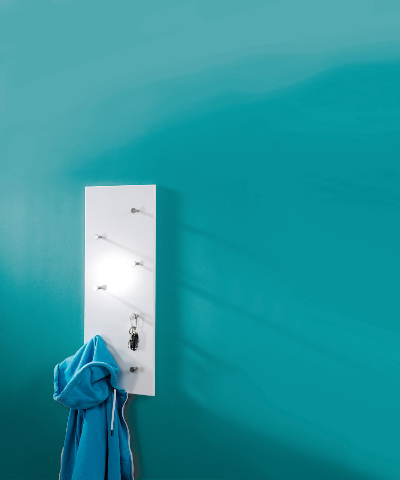 Weiße Wandgarderobe mit mehreren Haken, an der ein blauer Hoodie und ein Schlüsselbund hängen, vor einer türkisfarbenen Wand. Perspektive von vorne.