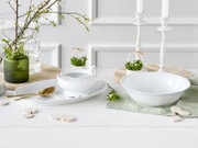 Weiße Porzellan-Servierschale mit zartem Reliefdekor auf hellem Tisch, daneben passende Platte und Sauciere, dekoriert mit Frühlingszweigen und Kerzenständern; Perspektive: leicht erhöhte Frontansicht.