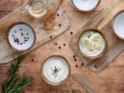 Draufsicht auf rustikale Keramikschalen mit Salz und bunten Pfefferkörnern, geschnittenem Fenchel und cremigem Dip mit Rosmarin auf Holzbrettchen, daneben Öl im Glas und Holzlöffel auf Holztisch