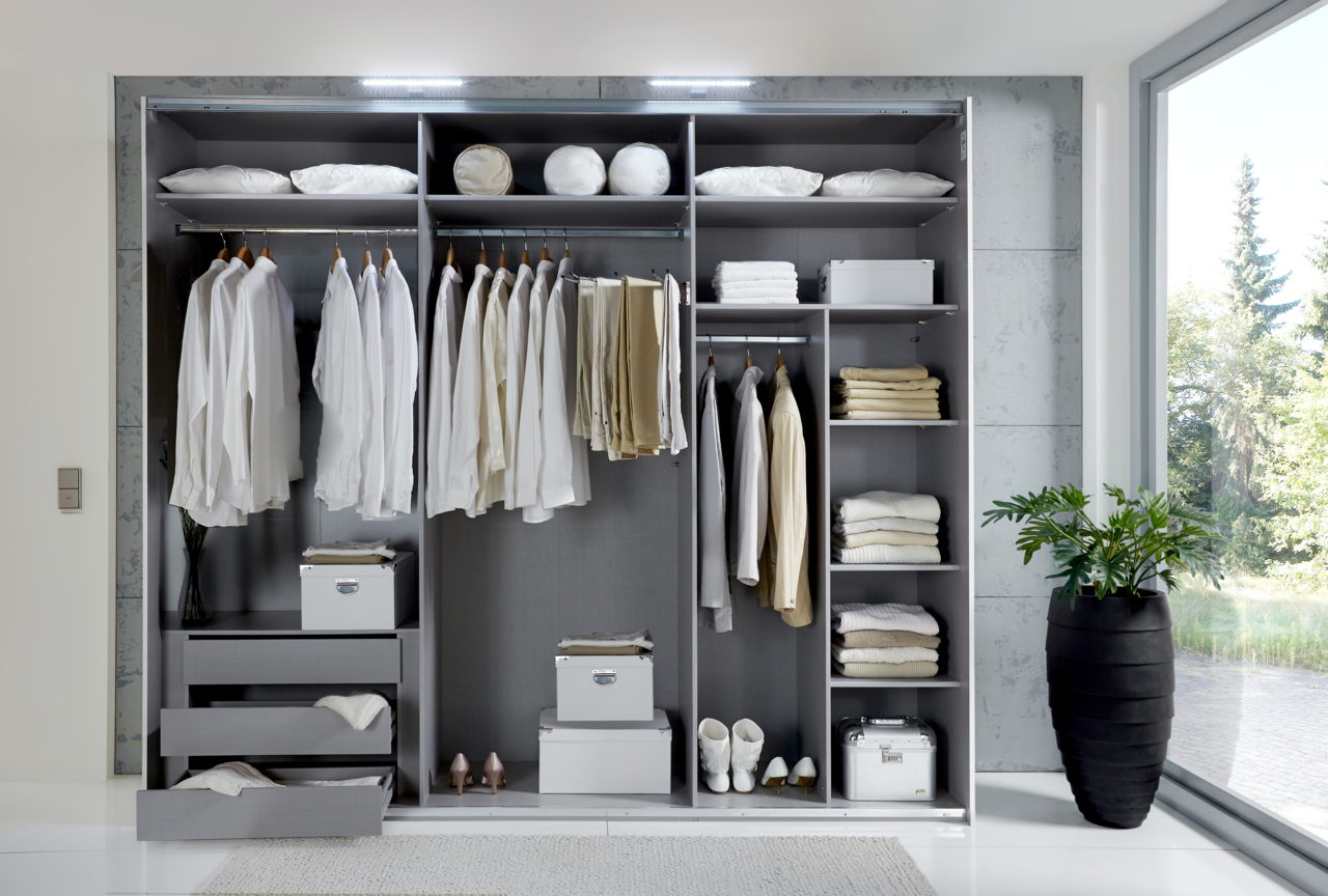 Offener Schwebetürenschrank mit drei Türen, gefüllt mit Kleidung, Kissen und Accessoires, von vorne betrachtet.